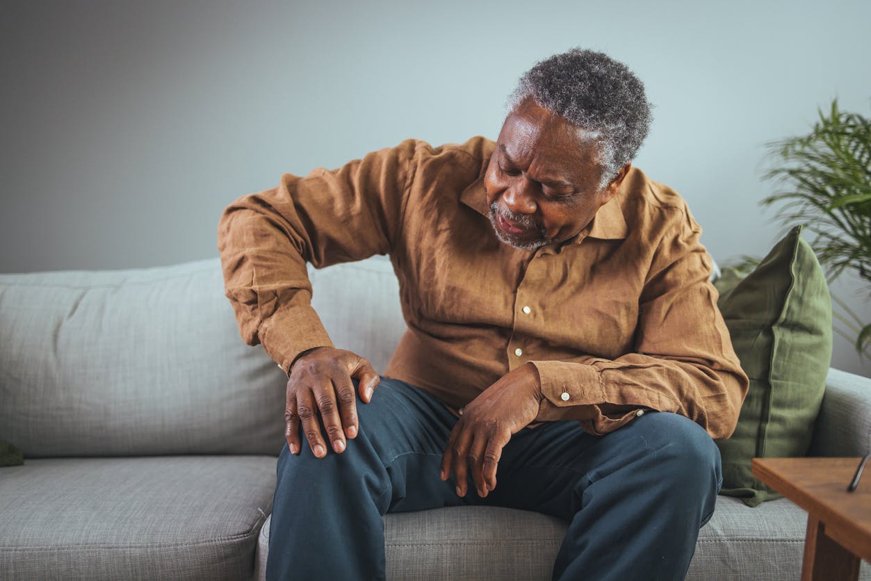 senior african american man feeling pain in his knees from arthritis while on a couch stock photo